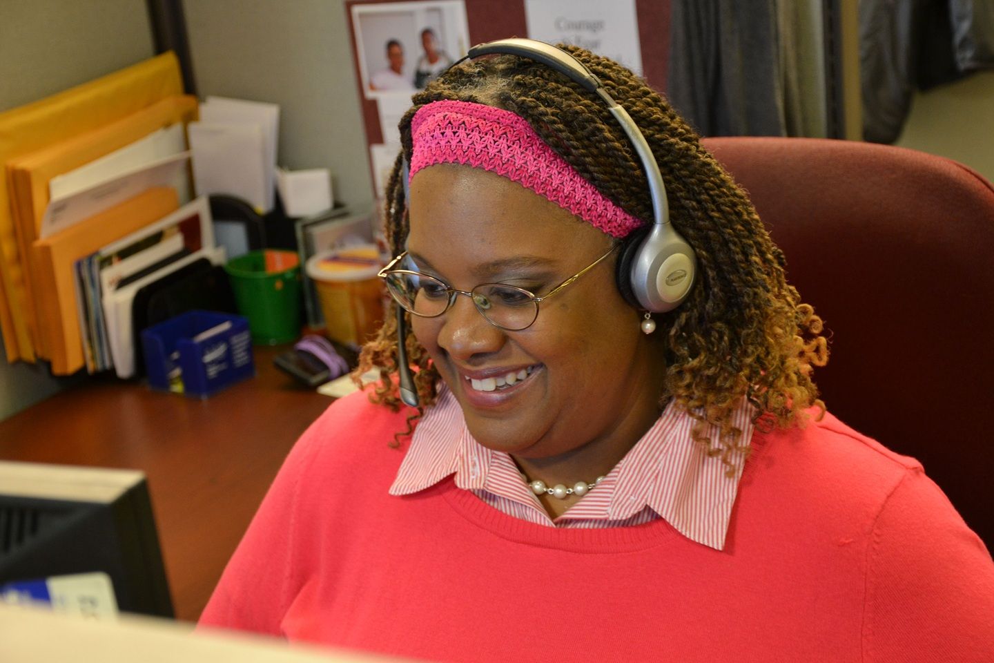A woman with glasses and headphones smiles as she looks down to her desk.