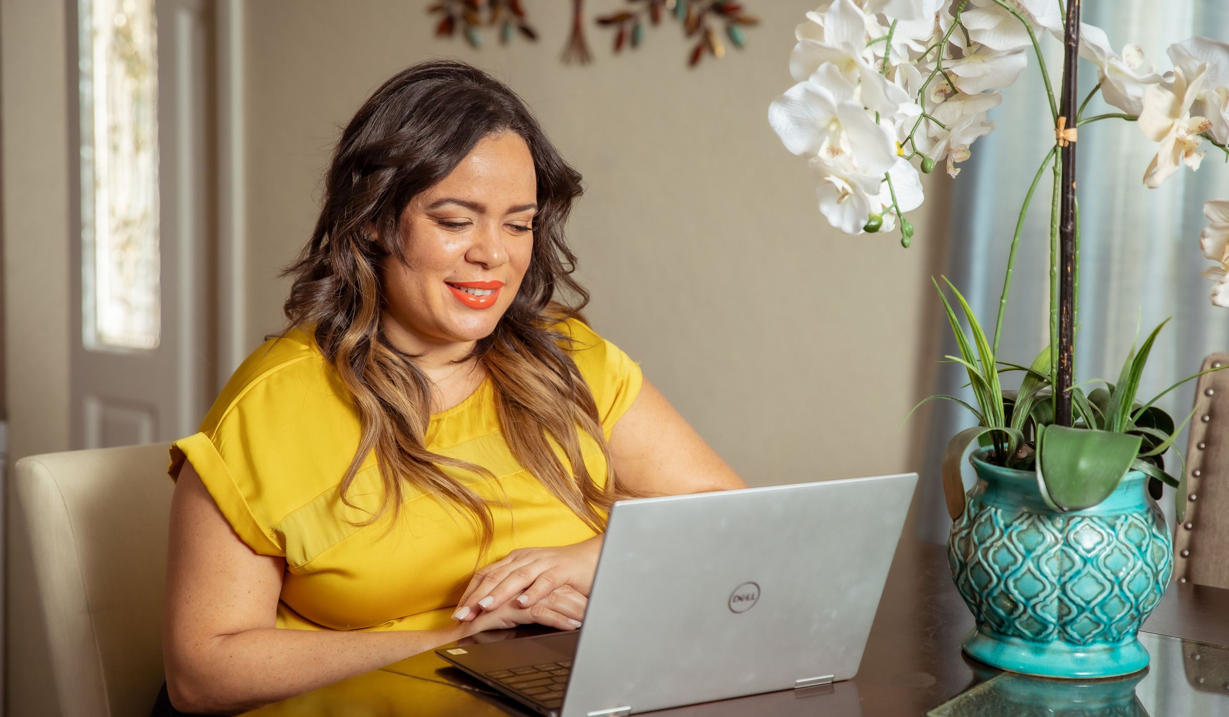 Una mujer con blusa amarilla sonríe ante una computadora portátil junto a una orquídea.