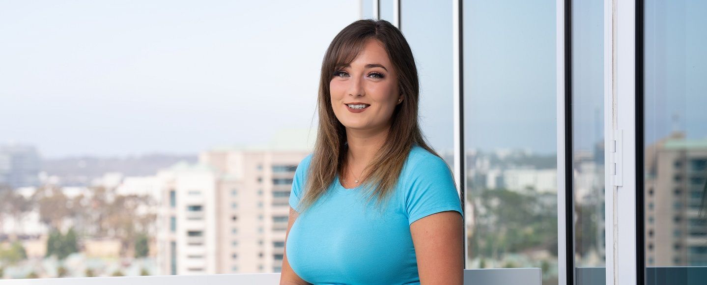 A woman in a light blue shirt smiles in front of a large window with a cityscape view.