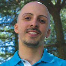 A person with a goatee wearing a blue collared shirt stand outside and smiles. 
